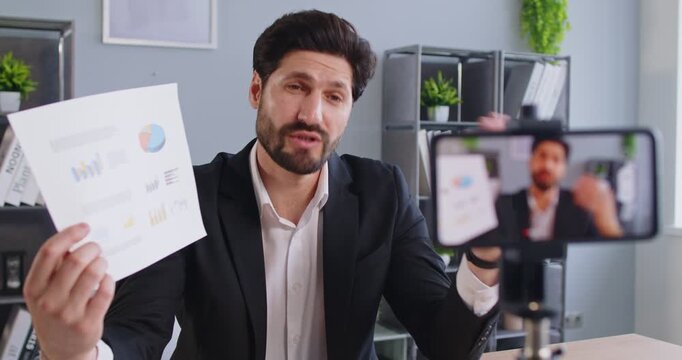Young man sitting in office having online webinar or video call talking about company finance. Confident male blogger having online business e-learning recording video blog on mobile phone. 4k video.
