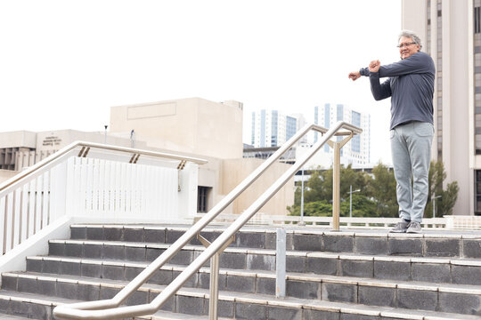 Mature man stretching arms across chest on plaza steps near metal rails in sportswear, copy space