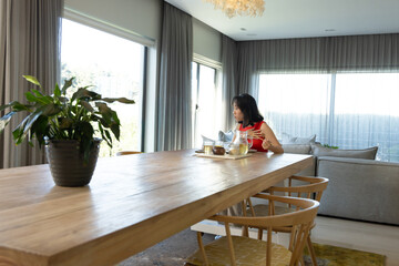 Fototapeta premium Asian woman wearing red top sitting at home dining table arranging tray with cups, copy space