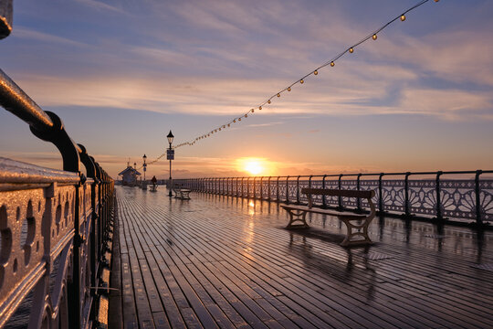 Sunrise blazes at the end of Penarth Pier, warm orange reflected in the wet decking as benches and ornate Victorian lamp posts line the rain-washed walkway.