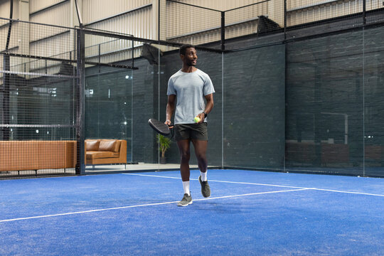 African American male in sportswear standing on blue padel court holding racket, ball under lights
