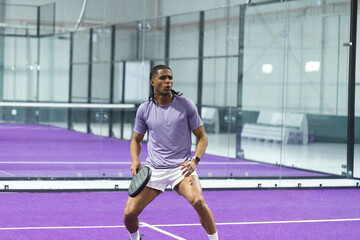Obraz premium African American male athlete in sportswear holding black paddle racket and moving on purple court