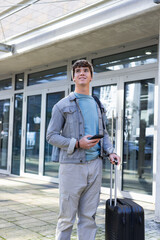 Man standing at glass doors wearing denim jacket and backpack holding smartphone, wheeled suitcase © wavebreak3