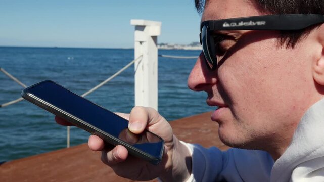 Man using voice assistant on smartphone at the beach, mobile technology, digital assistant, tech lifestyle, ocean view.