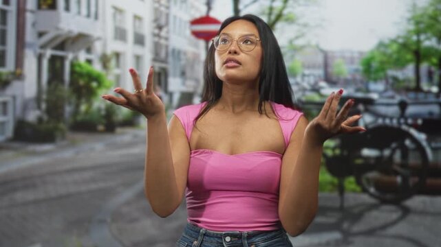 Young hispanic woman with hands on hips wearing pink top and glasses, bare shoulders visible, looking up on a street lined with buildings and bicycles; confidence assertiveness poise.
