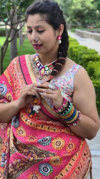 Indian woman in vibrant chaniya choli adjusting braided hair with kodi shells and fabric bangles, dressed in traditional Navratri garba outfit, showcasing colorful ethnic style outdoors.