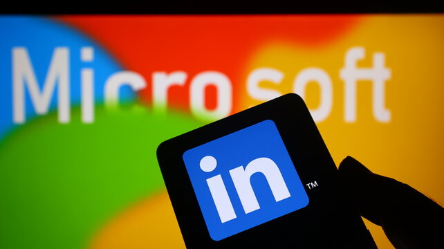 A hand holding a smartphone with the LinkedIn logo in front of a Microsoft logo background