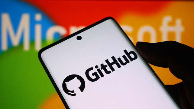 A hand holding a smartphone with the GitHub logo on the screen in front of a Microsoft background