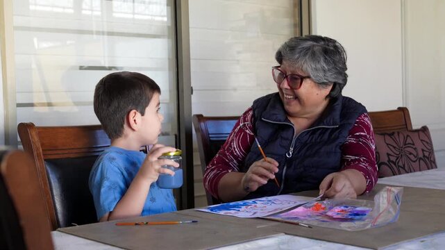 A cheerful grandmother and her young grandson share a joyful moment painting with bright watercolors on a patio. Authentic smiles beautifully highlight strong intergenerational bonding and creativity.