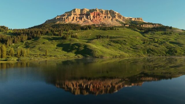 Beartooth Highway in Montana scenic view featuring majestic red rock mountain reflected in calm alpine lake waters surrounded by lush green meadows and pine forests under clear blue sky.
