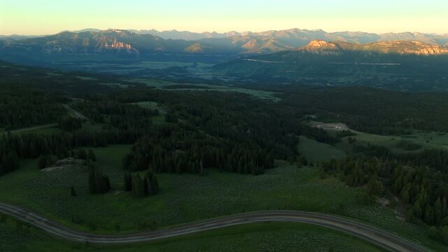 Aerial footage of Beartooth Highway in Montana reveals stunning mountain panorama with winding road through lush green forests and valleys bathed in warm sunrise light.