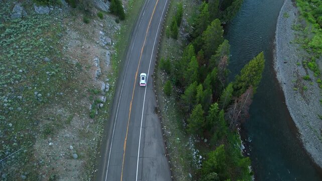 Drone footage follows a white car traveling on a winding mountain highway beside a turquoise river. Evergreen pine trees line the scenic route through rugged wilderness terrain.
