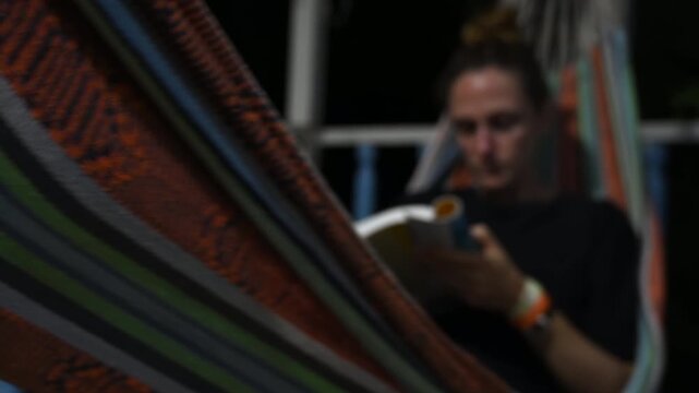 woman reading a book at night while gently swinging on a hammock on a house terrace. Soft lighting and calm atmosphere create a relaxing and intimate scene, ideal for concepts of leisure, solitude