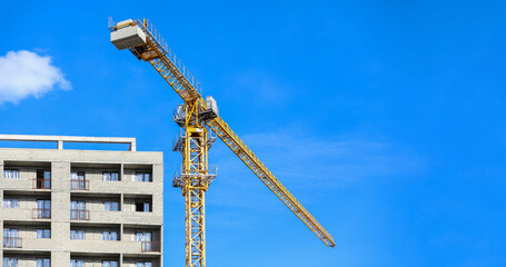 Yellow construction tower crane working on a residential building development, building new homes and modern architecture against a bright blue sky, developing urban infrastructure © Newleks