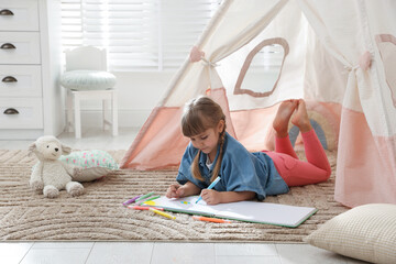 Little girl drawing in sketchbook near toy wigwam at home © New Africa