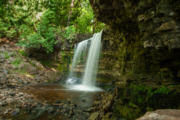 Obraz premium A long-exposure of a majestic waterfall cascading over a moss-covered cliff into a rocky pool. Surrounded by lush forest greenery and serene, peaceful natural scenery ideal for outdoor escape.