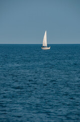 Fototapeta premium A lone white sailboat glides across a calm blue sea beneath a clear sky, highlighting open horizon, peaceful solitude and minimalist composition for a relaxed, nautical and leisurely summertime scene.
