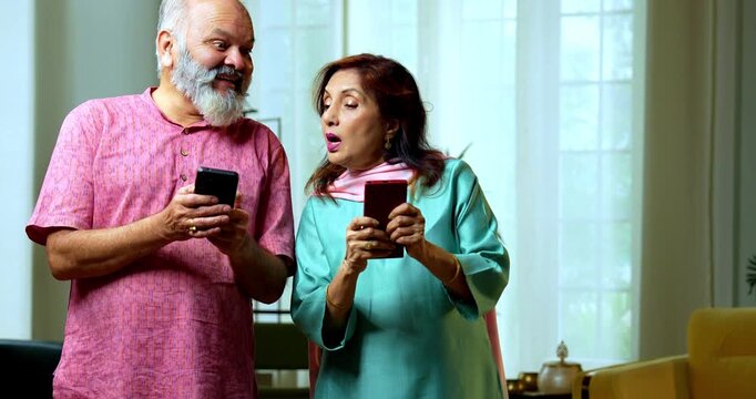An Indian senior couple standing side by side and typing on their smartphones. They are playfully peeping into each other screens with curious and funny expressions in their living room