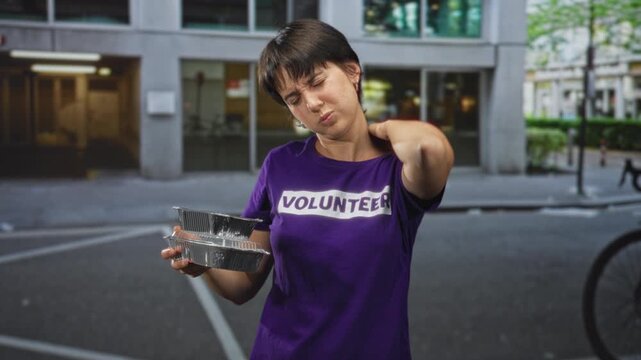 Woman in purple volunteer shirt holding stacked aluminum takeout containers rubs neck, shows neckache and winces on urban street; fatigue volunteering.