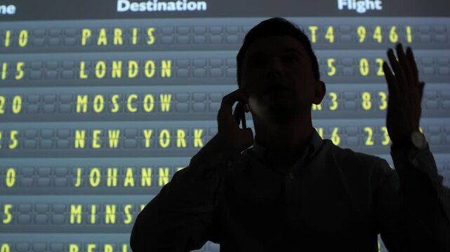 Silhouette of a man talking on a mobile phone, standing in front of a split flap display showing flight information and international destinations at an airport terminal