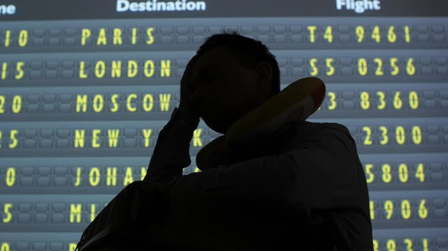 Silhouette of a traveler with a neck pillow holding head in hands, expressing frustration due to a cancelled or delayed flight shown on an airport departure board