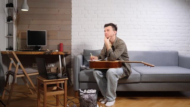 Young man sitting on a sofa with an acoustic guitar, covering his face with hands, surrounded by crumpled papers in a bin and on the floor, indicating stress and artistic struggle