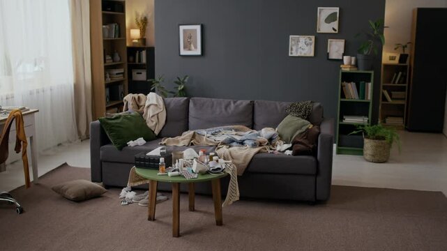 Wide establishing shot of domestic interior of sick persons home, messy couch with pile of crumpled blankets and pillows, table with medicines, pills, tissues, blood pressure monitor device at daytime
