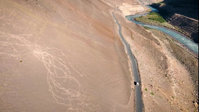 Cinematic drone tracking shot following a 4x4 SUV driving on a remote dirt road toward massive Himalayan mountains.