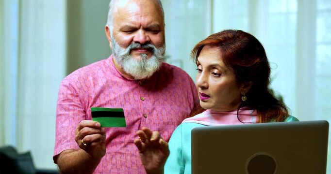 Senior Indian couple standing in a modern living room, using a debit card for an online transaction and showing it to the camera with confidence and smiles
