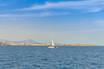 Fototapeta premium Catamaran with sail in water of Bahia Alicante bay Mediterranean Sea and Costa Blanca White coast, Cabo de la Huerta cape in Alicante city, mountain range background, Valencian Community, Spain