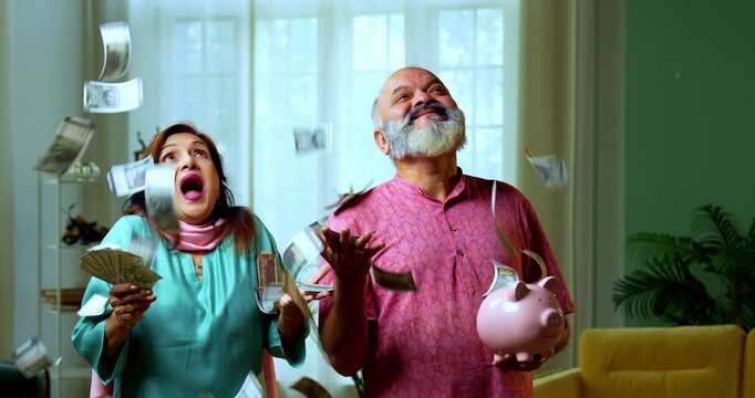 Senior Indian couple holding a five hundred rupee money fan and a piggy bank, looking surprised as cash is raining from above, denoting wealth growth and profits