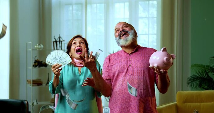 Senior Indian couple holding a five hundred rupee money fan and a piggy bank, looking surprised as cash is raining from above, denoting wealth growth and profits