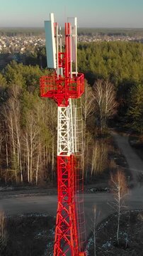 Close up drone shot of a red and white metal lattice mast with transmitters. Telecommunication infrastructure located near a forest and village under clear sky at sunset