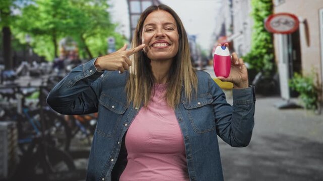 Woman points finger to cheek while holding a pink toothpaste dispenser on street near bicycles and storefronts; cheerful dental care.