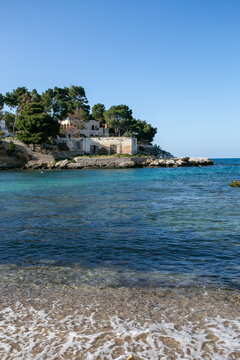 A Mediterranean-style coastal scene featuring a small structure on a rocky shoreline surrounded by trees, with clear blue water gently meeting the shore. The setting suggests a secluded cove or bay ty