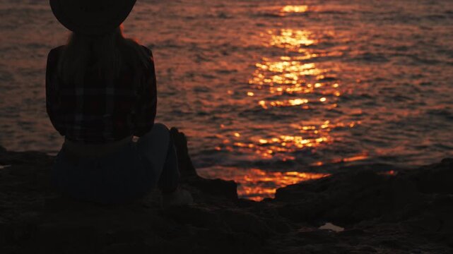 Silhouette of lone woman in cowboy hat sit on rocky cliffs, admiring golden sunset over calm ocean, evoking freedom, reflection, adventurous American spirit.