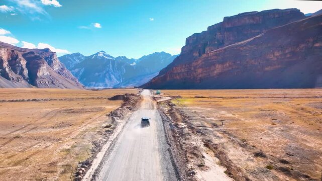 Cinematic drone tracking shot following a 4x4 SUV driving on a remote dirt road toward massive Himalayan mountains in spiti valley