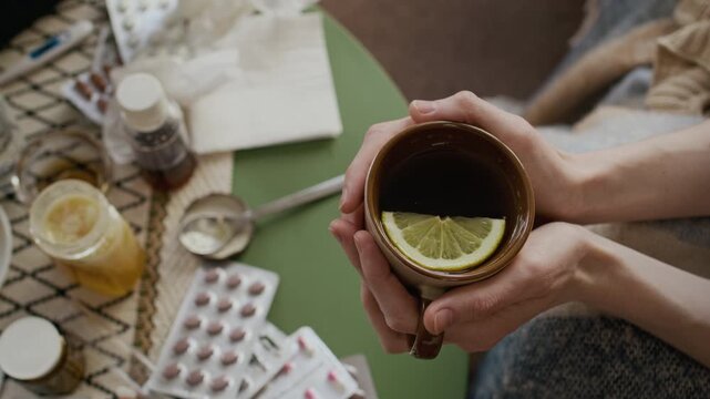 Top view close-up of hands of anonymous female flu or seasonal cold sufferer holding cup of hot tea with lemon and honey, while combining pharmaceutical prescription medicines and traditional remedies