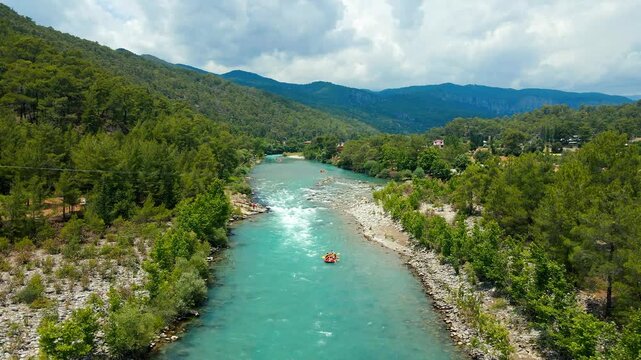 Aerial view of turquoise river winding through lush green hills. Clouds drift above the mountains' peaks. Rapid waters flow through rocky, forested banks. Vibrant foliage lines both riverbanks