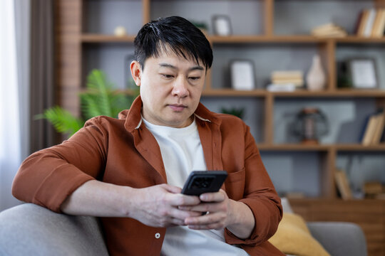Asian man sitting comfortably on a sofa, looking intently at his smartphone, connecting, communicating, and interacting with technology in his modern home environment