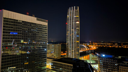 Fototapeta premium Modern city buildings with glass facade and tall skyscraper. Wide shot of office blocks reflecting night lights with a residential tower.