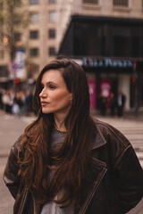 Obraz premium Candid profile portrait of a stylish young woman with long, rich brown flowing hair and a thoughtful, serious facial expression, looking away on a blurred city street with buildings, traffic