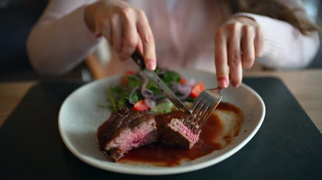 Close up of woman cutting juicy grilled steak served with rich sauce and fresh salad