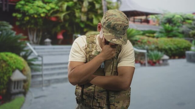 Man in army uniform with hand covering mouth and crossed arms in front of building entrance; contemplation duty.