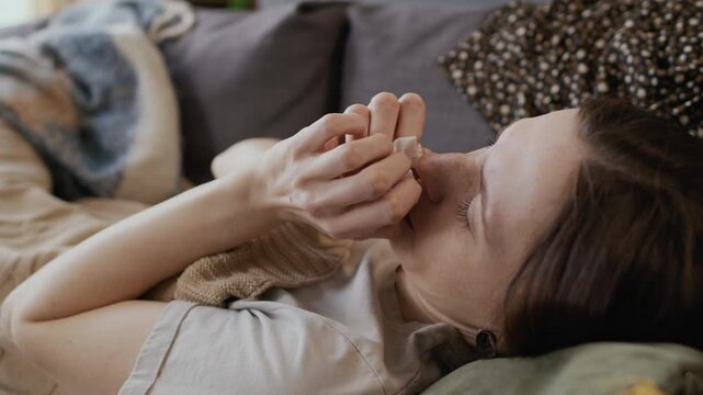 Medium side shot of Caucasian girl with bleary red eyes, suffering from seasonal allergy lying on couch, sneezing, coughing while clutching tissue in hand, wiping congested nose