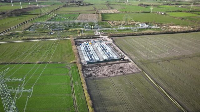 Aerial footage of numerous containerized battery units arranged in orderly rows, integrated with a nearby electrical substation and high-voltage transmission lines. Grid-scale battery storage.
