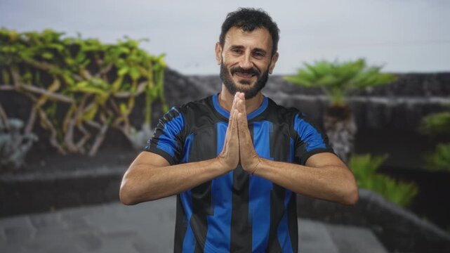 Man with hands pressed in namaste gesture wearing blue black soccer uniform on a building rooftop, smiling with visible beard and palms together; calm gratitude.