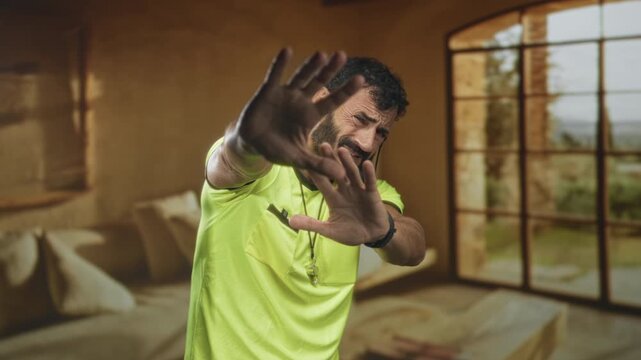Referee man with whistle holds hands up blocking face in building interior by large window and cushions, neon yellow shirt visible; defensive.