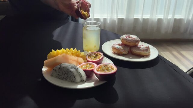 Tropical freshness and vitamins: woman scoops passion fruit pulp, mixes drink with spoon. Mango, papaya, jackfruit and donuts on buffet table. 