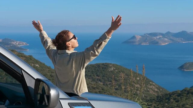 Woman raising arms in freedom admiring sea view. Happy woman on a road trip standing through the sunroof of a car with her arms outstretched, enjoying the breathtaking view of the sea with islands